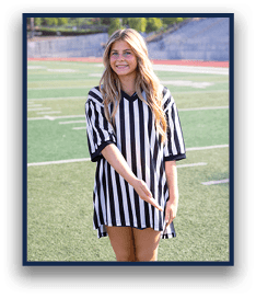A person with long hair wearing a black and white striped referee shirt is standing on a sports field smiling and gesturing with one hand. Description generated by AI