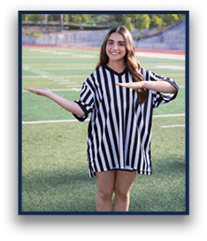A person with long hair wearing a black and white striped shirt is standing on a sports field smiling and holding their arms out horizontally at different heights. Description generated by AI