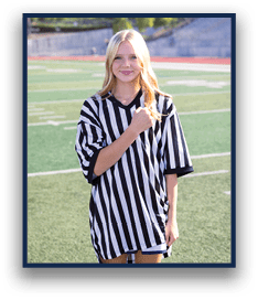A person with long hair wearing a black and white striped referee shirt stands on a sports field with a slight smile and one hand raised to their chest. Description generated by AI