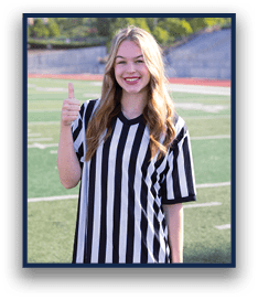 A person with long hair wearing a black and white striped shirt is standing on a sports field smiling and giving a thumbs up. Description generated by AI