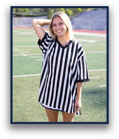 A person with long light hair is smiling and standing on a sports field wearing a black and white vertically striped oversized shirt with one arm raised behind their head. Description generated by AI