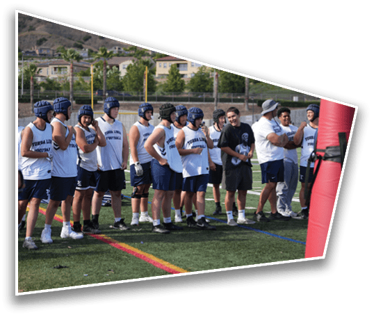 A group of people wearing sports helmets and sleeveless shirts stand on a field near a large red training dummy, appearing to prepare for a football practice. Description generated by AI