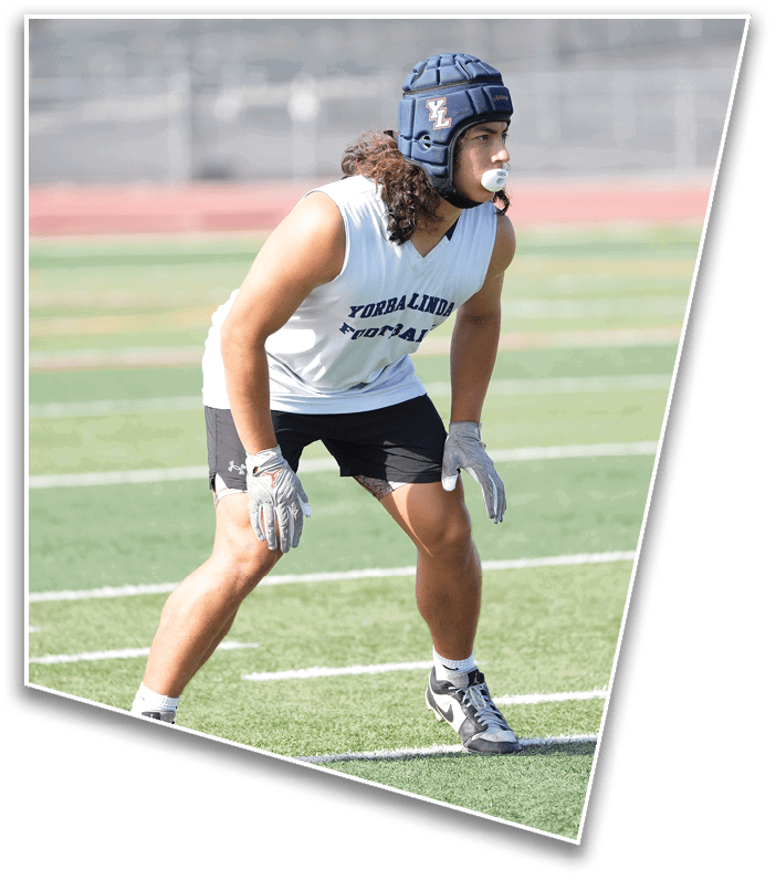 A person wearing a sleeveless sports shirt, shorts, gloves, cleats, and a padded helmet is crouched in an athletic stance on a football field. Description generated by AI