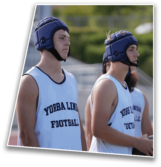 Two people wearing blue padded helmets and white sleeveless shirts with “YORBA LINDA FOOTBALL" printed on them stand outdoors near a sports field. Description generated by AI