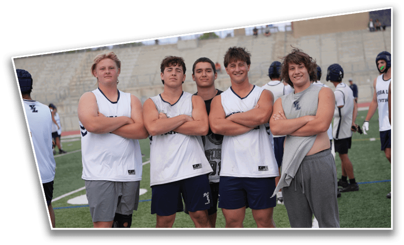 Five people wearing athletic clothing stand on a sports field with their arms crossed, smiling, while others in similar attire are visible in the background. Description generated by AI