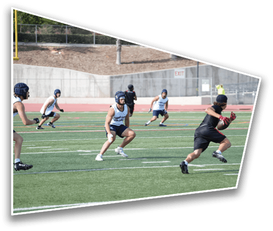 People wearing helmets and athletic clothing are playing a sport on a green field with a track and concrete wall in the background. Description generated by AI