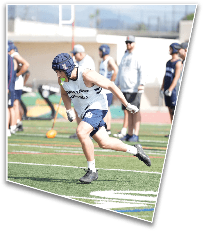 A person wearing athletic gear and a protective helmet is running on a sports field while several people stand in the background watching. Description generated by AI