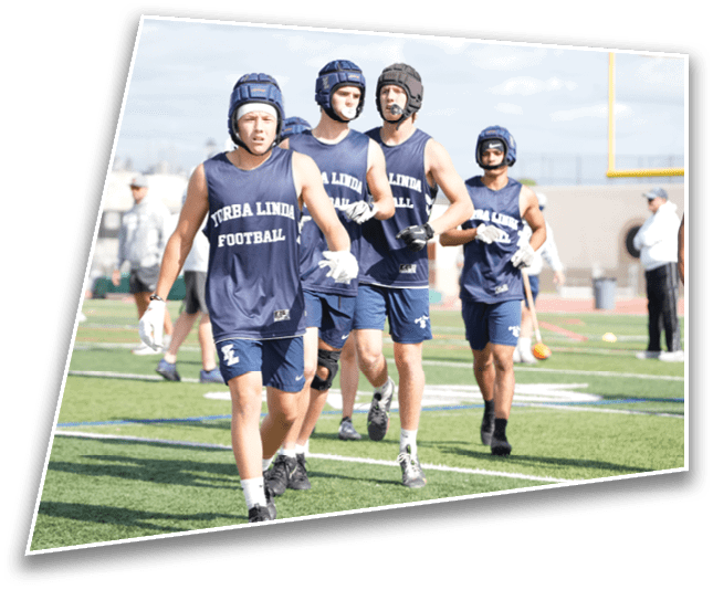 Four people wearing blue sports helmets and matching sleeveless shirts with “TURBA LINDA FOOTBALL" printed on them are walking on a sports field. Description generated by AI