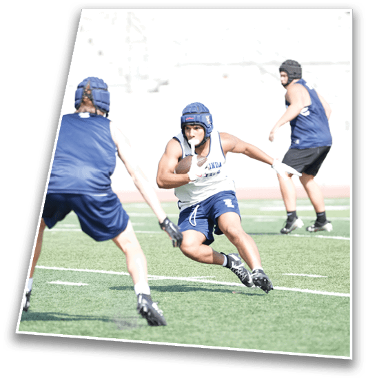 Three people wearing helmets and athletic gear are playing a sport on a grassy field, with one person running while holding a ball and the others positioned defensively. Description generated by AI
