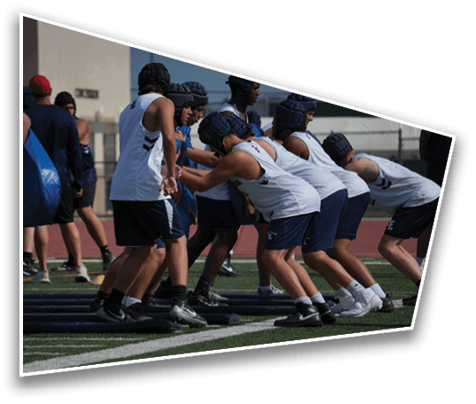 People wearing sports helmets and athletic clothing practice blocking drills on a football field with coaches holding tackling pads. Description generated by AI