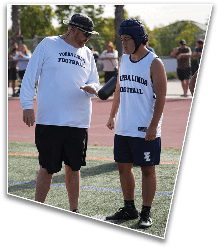 Two people wearing Yorba Linda Football shirts are standing on a sports field with one person appearing to give instructions to the other who is wearing a padded helmet. Description generated by AI