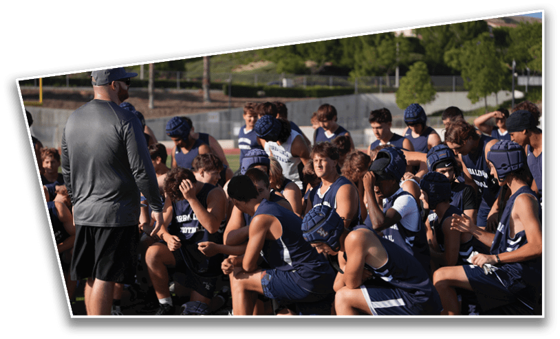 A group of people wearing sports uniforms and helmets are gathered outdoors on a field, some sitting and some standing, while one person in a long-sleeve shirt and shorts appears to be addressing them. Description generated by AI