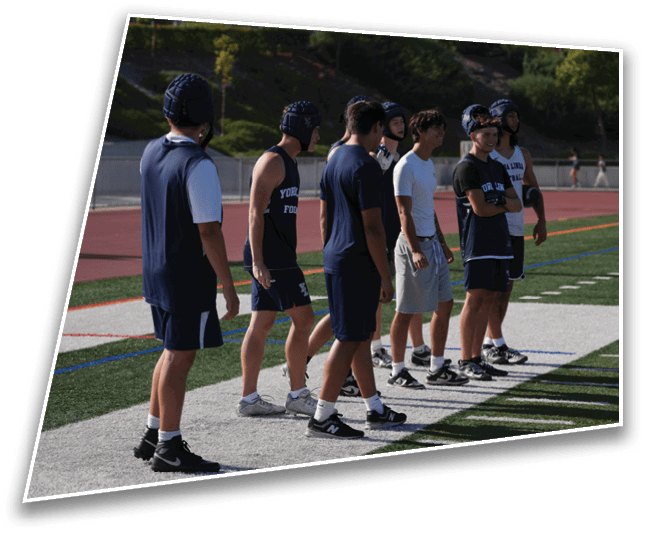 A group of people wearing athletic clothing and helmets stand in a line on the edge of a sports field with trees and a track in the background. Description generated by AI