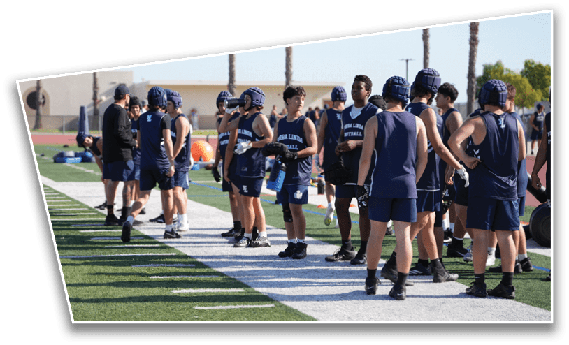 A group of people wearing athletic gear and helmets stand on the sidelines of a sports field with palm trees and buildings in the background. Description generated by AI