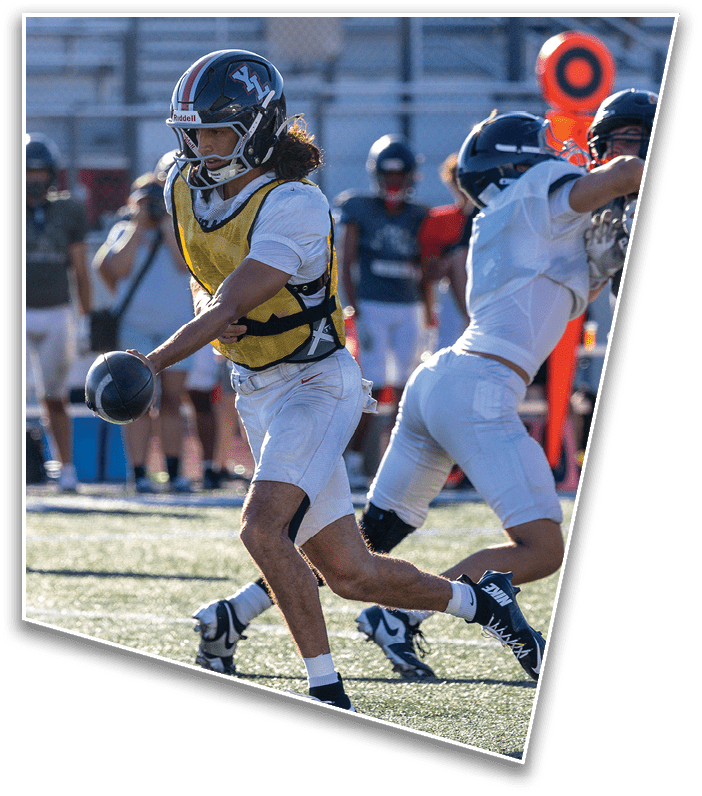 A person wearing a football helmet and a yellow practice vest is on a field with blurred bleachers and other players in the background. Description generated by AI