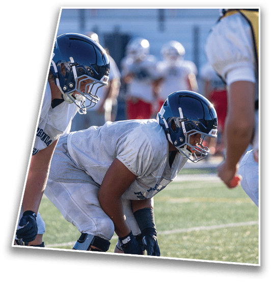 Two people wearing football helmets and white jerseys are positioned closely on a field with blurred figures in the background. Description generated by AI