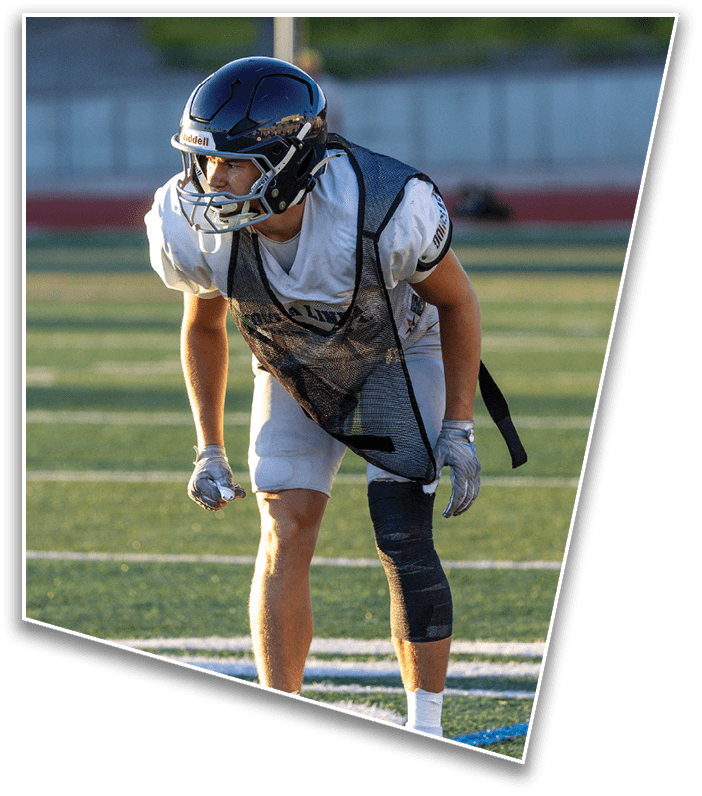 An adult wearing a football helmet, gloves, and a black mesh practice jersey is crouched on a grassy field, appearing ready to move. Description generated by AI