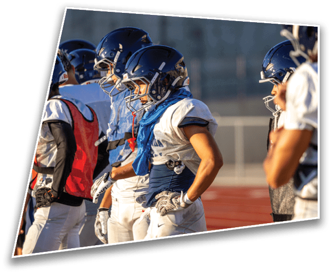 Several people wearing football helmets and uniforms stand closely together on a sports field, appearing focused and ready for action. Description generated by AI