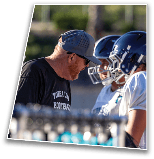An adult wearing a cap and sunglasses is leaning forward and speaking to two people in football helmets and uniforms during an outdoor practice or game. Description generated by AI