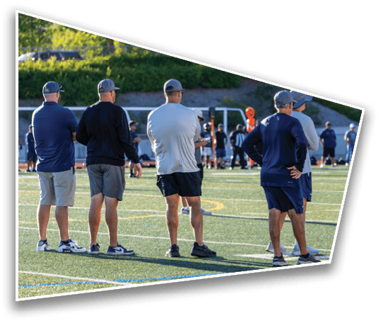 Several adults wearing casual athletic clothing and caps stand on the sidelines of a sports field with other people and equipment visible in the background. Description generated by AI