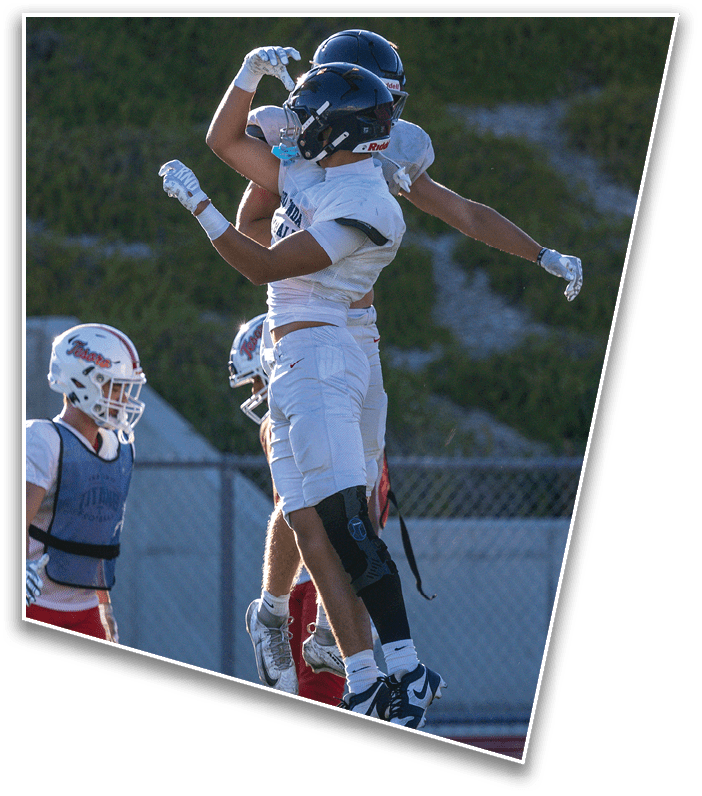 Two people in football uniforms and helmets are jumping and celebrating on a field with a fence and greenery in the background. Description generated by AI