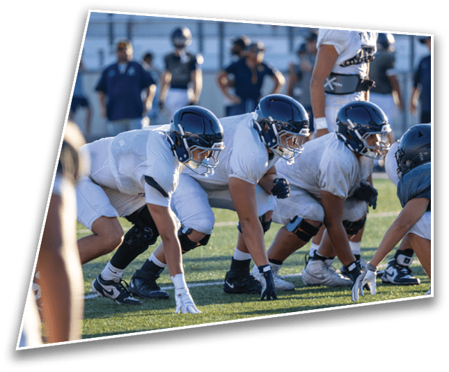 Three people in football helmets and white practice uniforms are crouched in a ready position on a grassy field during a practice session. Description generated by AI