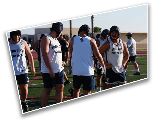 People wearing sleeveless sports jerseys and protective headgear are standing and interacting on a football field during daylight. Description generated by AI