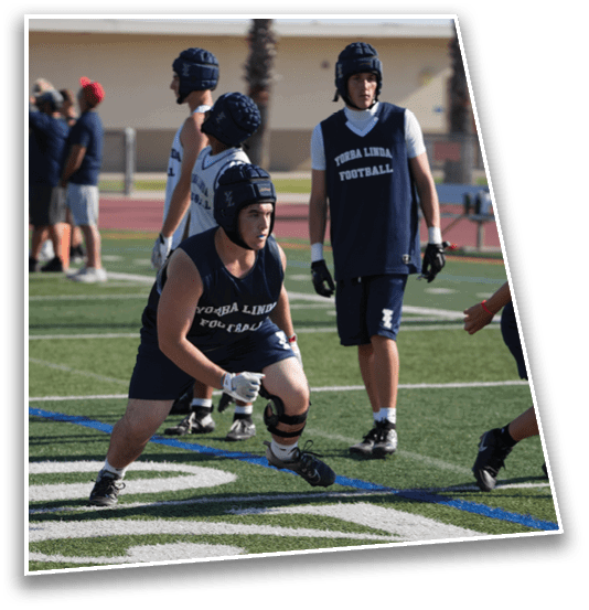 A person wearing a helmet and knee brace is crouched in an athletic stance on a football field while others in matching sports uniforms stand nearby. Description generated by AI