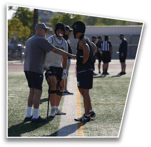 An adult wearing a cap and casual sportswear is pointing and talking to two people in sports gear and helmets on a field with others in the background. Description generated by AI