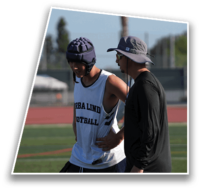 Two people stand on a sports field, one wearing a padded helmet and sleeveless shirt with text, and the other wearing a hat and long-sleeve shirt, appearing to converse or coach. Description generated by AI