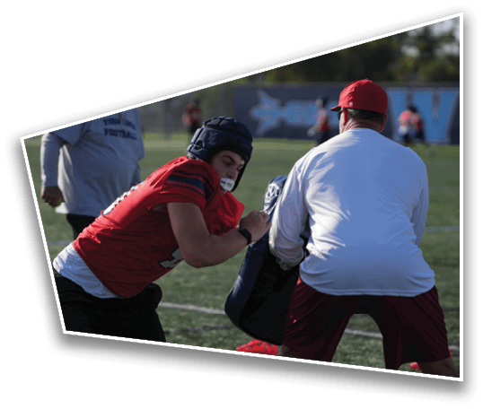 A person wearing a red sports jersey and helmet is practicing football tackling on a field with another person holding a padded blocking shield. Description generated by AI