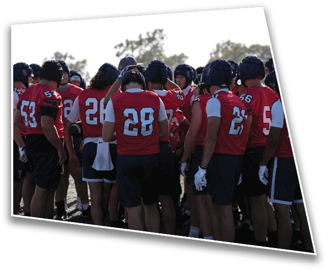 A group of people wearing red sports jerseys and helmets are gathered closely together on a grassy field, appearing to be in a team huddle or discussion. Description generated by AI