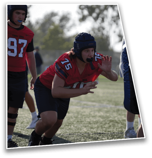 A person wearing a red sports jersey and protective headgear is crouched in an athletic stance on a field while another person stands nearby. Description generated by AI