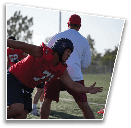 An adult wearing a red sports jersey and helmet is leaning forward in an athletic stance on a field while another adult in a white shirt and red cap stands nearby. Description generated by AI
