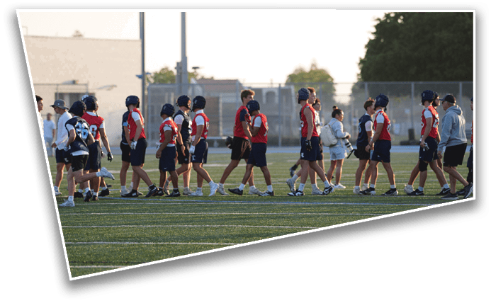 People wearing sports uniforms and helmets are walking on a field, likely after a game, with some adults and a fence in the background. Description generated by AI