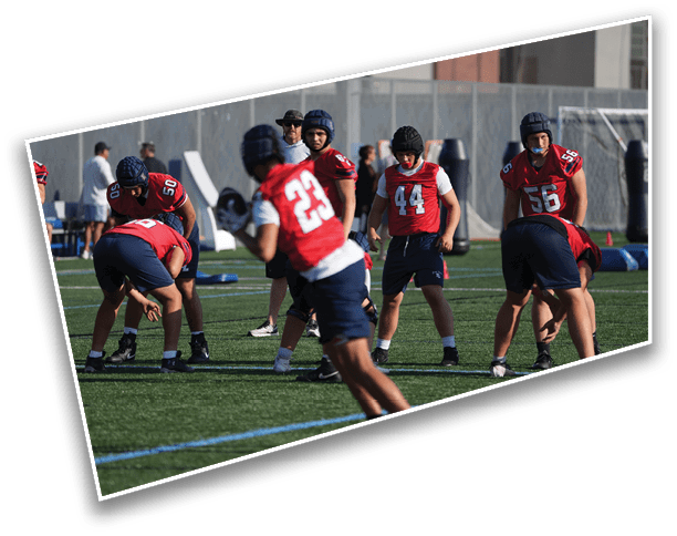 People wearing red sports jerseys and helmets are practicing football drills on a green field with a coach observing in the background. Description generated by AI