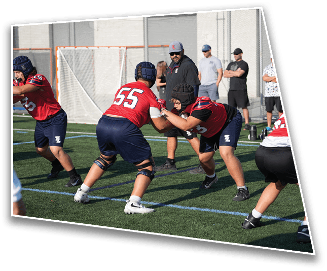 People wearing red and blue sports uniforms and helmets are practicing football drills on a green field with a goal net and several people watching in the background. Description generated by AI