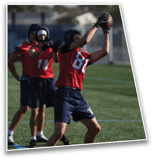 Three people wearing red sports jerseys and helmets are playing football on a grassy field, with one person catching a football while the others watch. Description generated by AI