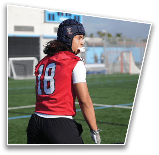 A person wearing a red sports jersey with the number 18, a protective helmet, and a mouthguard is standing on a sports field looking to the side. Description generated by AI