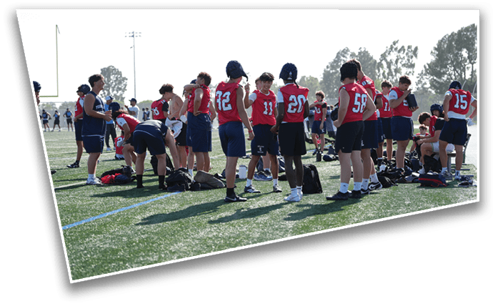 A group of people wearing red and blue sports jerseys are gathered on a grassy field, some standing and some bending over near bags, with trees and other people visible in the background. Description generated by AI