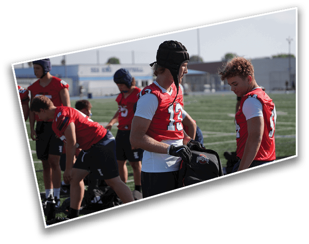 Several people wearing red sports jerseys and protective headgear are gathered on a grassy field, preparing for an activity. Description generated by AI
