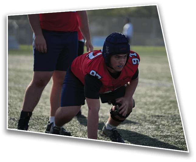A person wearing a red sports jersey and protective headgear is crouched on a grassy field in a ready position with another person standing nearby. Description generated by AI