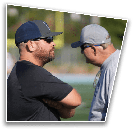 Two adults wearing baseball caps and sunglasses are engaged in a conversation outdoors on a sports field. Description generated by AI