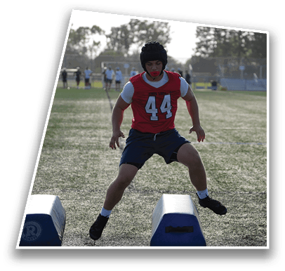 A person wearing a red sports jersey and helmet is jumping over training pads on a grassy field with other people and trees in the background. Description generated by AI