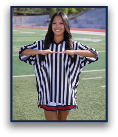 A person with long hair wearing a black and white striped referee shirt is standing on a sports field smiling and holding their hands parallel in front of them. Description generated by AI