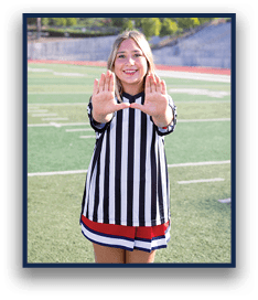 A person wearing a black and white striped referee shirt and a red, white, and blue skirt is standing on a sports field smiling and holding their hands up with palms facing forward. Description generated by AI