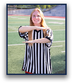 A person wearing a black and white striped referee shirt is standing on a sports field making a timeout gesture with their arms. Description generated by AI