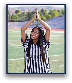 A person wearing a black and white striped referee shirt is standing on a sports field with hands pressed together above their head and smiling. Description generated by AI