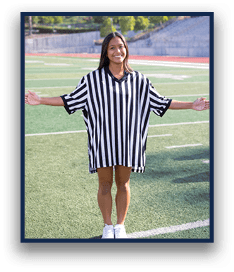 A person wearing a black and white striped referee shirt stands on a sports field with arms extended and smiling. Description generated by AI