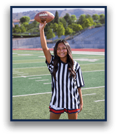 A person with long hair wearing a black and white striped shirt and red shorts is standing on a football field smiling and holding a football up in one hand. Description generated by AI